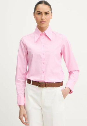 Donna che indossa una camicia rosa chiaro con bottoni infilata nei pantaloni bianchi con cintura marrone, posando con una mano in tasca.