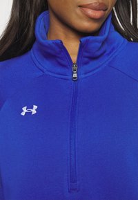 Sudadera atlética azul con alto cuello, cierre de cremallera y logo de Under Armour. Textura suave, diseño ajustado, con detalle de cremallera en contraste.