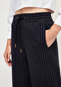 Marineblaue gestreifte Hosen mit Kordelzugbund, ausgestattet mit goldenen Akzenten, seitlichen Taschen und einer lockeren Passform. Glattes Gewebe mit vertikalen Linien.