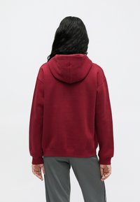 Person med langt svart hår iført en enkel rød hettegenser og grå bukser, stående vendt bort fra en enkel lys bakgrunn.