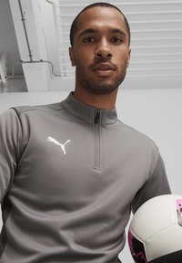 Grijze zip-up sporttrui met een gladde textuur, met een wit Puma-logo op de borst. De persoon houdt een voetbals met rozige accenten vast.
