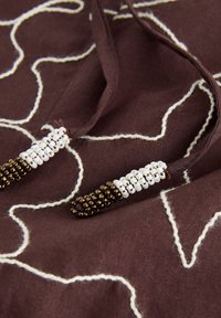 Tissu brun foncé avec des lignes blanches cousues et deux cordons ornés de perles blanches et bronze aux extrémités.