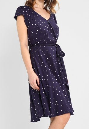 Kobieta w granatowej sukience w grochy, z krótkimi rękawami i wiązaniem w talii, pokazana od ramienia do kolan na jasnym tle.