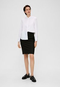 Witte overhem shirt met kraag, gecombineerd met een getailleerde zwarte rok en zwarte chunky loafers, tegen een effen achtergrond.