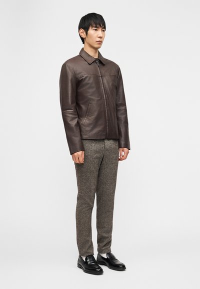 Άνδρας στέκεται φορώντας καφετί σκούρο δερμάτινο μπουφάν, γκρι υφασμάτινο παντελόνι, λευκές κάλτσες και μαύρα δερμάτινα μοκασίνια, μπροστά σε απλό φόντο.