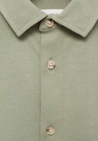 Ljusgrön polo skjorta tillverkad av texturerad tyg, med en krage och tre beige knappar längs knäppningen. Hållbara sömnadsdetaljer synliga.