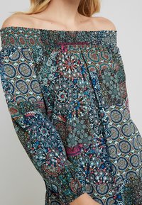 Olkapäältä avattu mekko, jossa on värikkäitä pyöreitä kuvioita, sinisiä, punaisia ja valkoisia yksityiskohtia. Valmistettu kevyestä, teksturoidusta kankaasta.