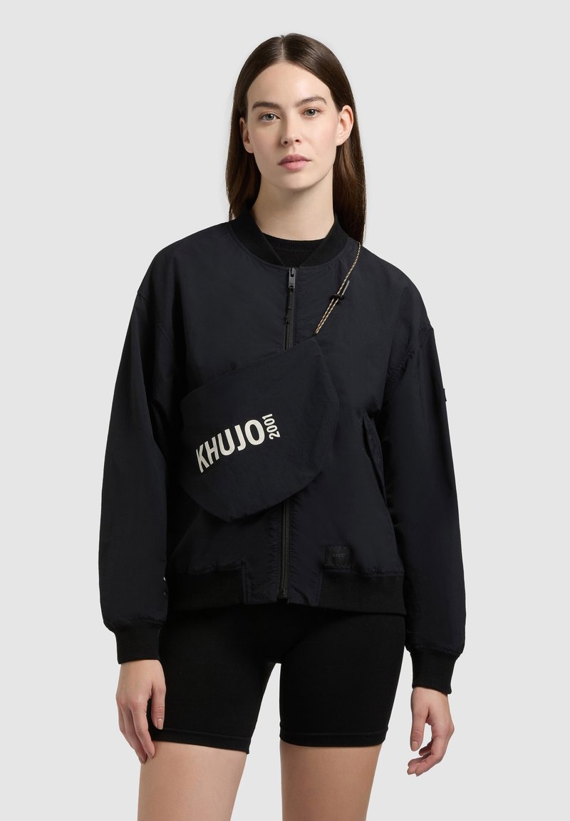 Jeune femme aux longs cheveux bruns portant une veste noire zippée, un short noir et un sac bandoulière avec l'inscription "KHUJO 2001".