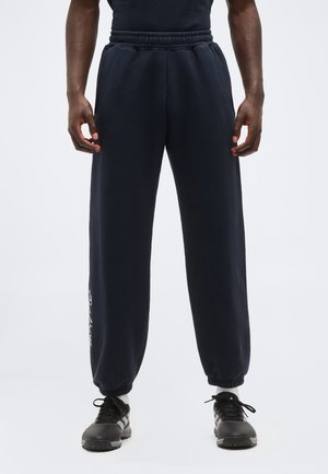 Homme portant un pantalon de jogging noir avec taille élastique et poignets élastiques, ainsi que des chaussures de sport noires, debout devant un fond uni.