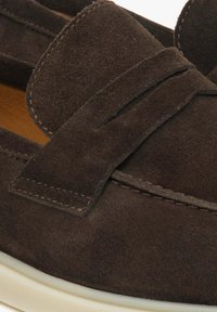 Dunkelbraune Wildleder-Loafers mit einem gesteppte Schleifendetail und einer kontrastierenden weißen Gummisohle. Strukturiertes Finish mit gesteppten Akzenten.