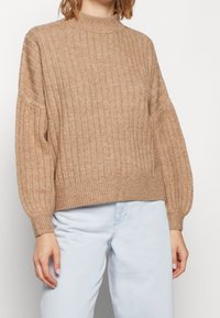 Beżowy, prążkowany sweter z wysokim kołnierzem i szerokimi rękawami, o miękkiej fakturze i krótkim kroju, zestawiony z jasnoniebieskimi jeansami.