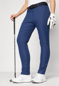 Modre športne hlače iz lahkega blaga s tesnim krojem, elastičnim pasom z dolgim trakom ter kombinirane z belimi golf čevlji in rokavico.