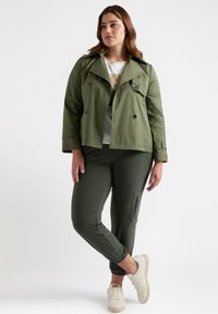 Olivgrön dubbelknäppt jacka med stora knappar, kombinerat med mörkgröna avsmalnande byxor med dragkedjefickor, och beige sneakers.