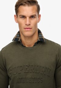 Brązowa bluza z wyszytym napisem "SUPERDRY ATHLETICS". Posiada kraciasty kołnierzyk w ciemnozielonym, niebieskim i beżowym kolorze, z wykończeniem z fakturowanej tkaniny.