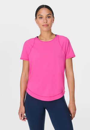 Vrouw met donker haar, gekleed in een roze sporttop met korte mouwen en donkerblauwe legging, staand tegen een effen witte achtergrond.