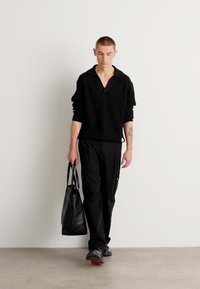 Schwarzer gerippter Pullover mit V-Ausschnitt, kombiniert mit schwarzen Cargo-Hosen. Das Modell trägt eine schwarze Tote-Tasche und hat dunkle Schuhe mit roten Akzenten an.