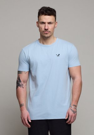 Mand med kort mørkt hår iført lyseblå t-shirt med lille krydset pagaj- og årerlogo, viser tatoveringer på begge arme, står neutralt.