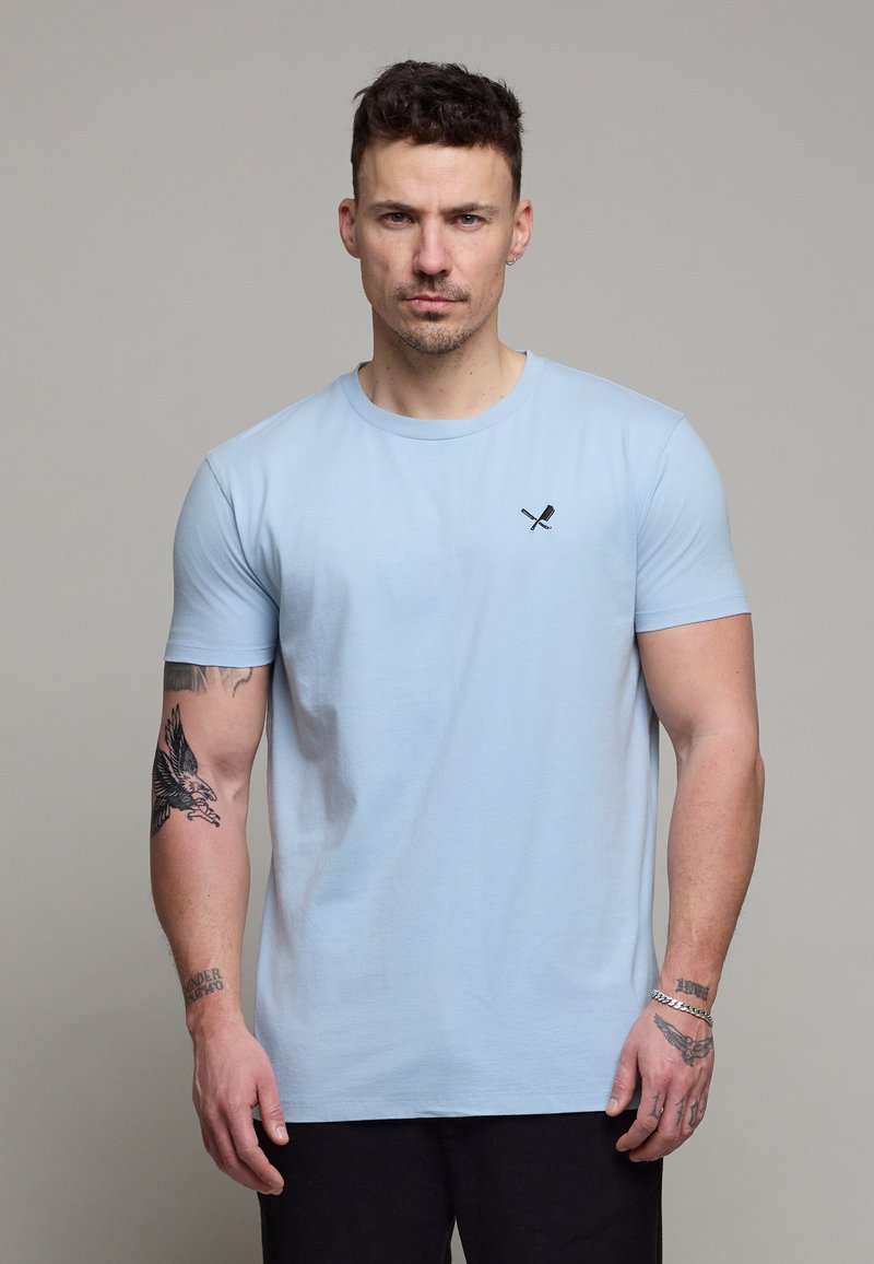 Homme aux cheveux courts et foncés portant un t-shirt bleu clair avec un petit logo de pagaies croisées, montrant des tatouages sur les deux bras, debout de façon neutre.