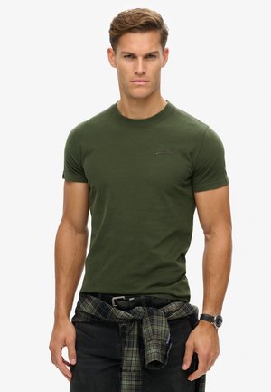 Dark green short-sleeve t-shirt with a crew neck and subtle logo detail. Worn with black jeans and a plaid shirt tied around the waist.
