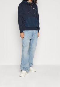 Felpa navy con coulisse regolabile, tasca kangaroo frontale, abbinata a jeans azzurri chiari e sneakers bianche. Vestibilità rilassata, texture morbida.