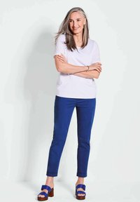 Witte t-shirt met korte mouwen, blauwe skinny jeans en blauwe platform sandalen met een bruine houten hak. Model staat met gekruiste armen en een glimlach.