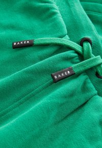 Vihreät fleece-housut, joissa on kiristysnyöri. Varustettu kahdella tarralla, joissa lukee "BAKER" valkoisella mustalla kankaalla. Teksturoitu materiaali, jossa on ribbireunat.