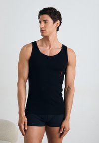 Schwarzes Baumwoll-Tanktop mit einem klassischen Rundhalsausschnitt, figurbetontem Design und einem kleinen roten Logo auf der linken Seite. Getragen mit passenden schwarzen Shorts.