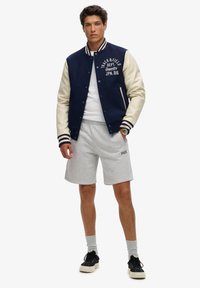 Chaqueta varsity en azul marino y crema con texto bordado, pantalones cortos de sudadera grises y zapatillas negras. El modelo muestra un look atlético casual.