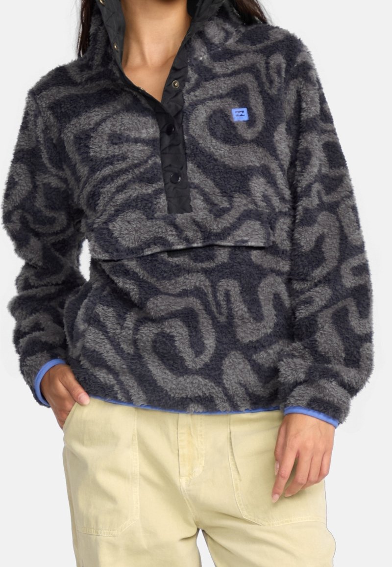 Femme portant un pull en polaire gris et noir à motif avec des boutons-pression noirs et un pantalon beige clair, mains dans les poches.