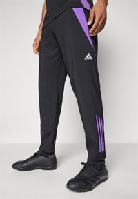 Zwarte sportbroek met paarse zijpanelen, voorzien van het Adidas-logo in het wit. Het materiaal lijkt glad met een taps toelopende pasvorm.