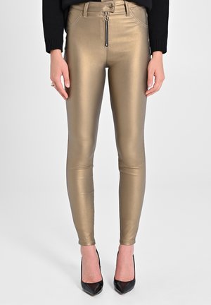 Obcisłe metaliczne złote spodnie z suwakiem z przodu, noszone z czarnymi szpilkami z ostrym noskiem i czarną bluzką z długim rękawem.