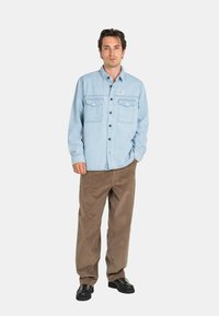 Hombre vestido con una camisa de botón a rayas azul claro, pantalones de pana marrones y zapatos negros, de pie contra un fondo claro y liso.