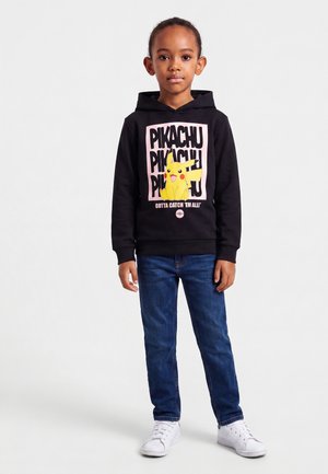 Enfant portant un sweat à capuche noir avec un Pikachu jaune et le texte "Gotta catch 'em all!", jean bleu et baskets blanches, debout devant un fond uni.