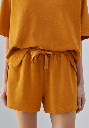 Person iført terry-cloth-shorts i sennepsgul med en matchende løs top, der viser snoretræk i taljen og sidelommer, stående med hænderne langs kroppen.