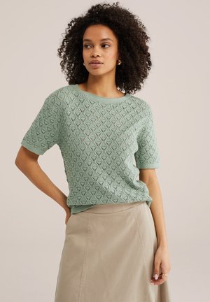 Frau mit lockigem Haar, die ein hellgrünes, gestricktes Kurzarmoberteil und einen beigen Rock trägt, vor einem einfarbigen Hintergrund stehend.