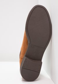 Zapato de ante marrón con punta redondeada y suela de goma texturizada. Incluye un tacón pronunciado y un marcador de tamaño grabado en la suela.