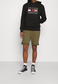 Felpa nera con cappuccio e dettaglio del logo, abbinata a pantaloni corti verde oliva. Indossata con scarpe alte nere e calzini bianchi alla caviglia.