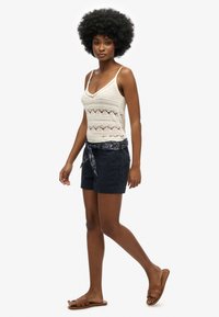 Top tank in crochet color crema, con motivi decorativi, abbinato a pantaloni corti blu navy con cintura a fiocco decorata. Sandali beige a fascia.