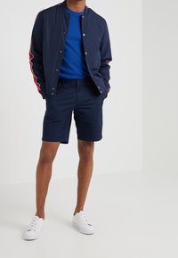 Muž v námořnické bomber bundě s červenými pruhy, modrém tričku, námořnických šortkách a bílých teniskách stojí s rukama v kapsách.