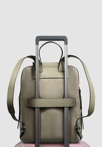 Sac à dos en cuir vert olive fixé à un chariot argenté, doté de deux poignées supérieures, d'une bandoulière amovible et d'un design minimaliste.