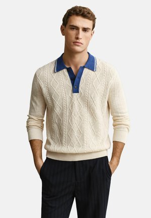 Man met handen in de zakken, gekleed in een crèmekleurige kabelgebreide trui met een blauwe kraag en donkere, fijn gestreepte broek, tegen een effen achtergrond.