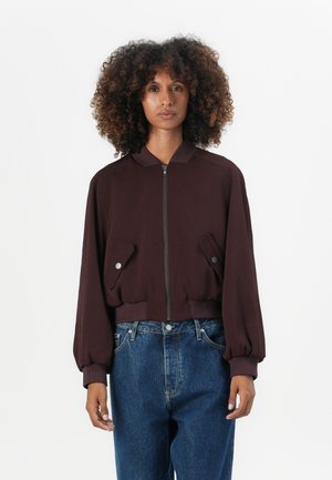 Burgunder bomberjakke med glidelås foran, to sidelommer med trykknapper, ribbede mansjetter og en cropped design. Båret over blå denimjeans.