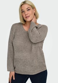 Blond žena s krátkými vlasy, která nosí zlaté kruhové náušnice a volný svetřík v barvě taupe, jednou rukou si sahá na krk.