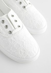 Weiße Leinenschuhe mit gestickten Blumenmustern, Metallösen und einer strukturierten Gummisohle. Abgerundete Zehenform.