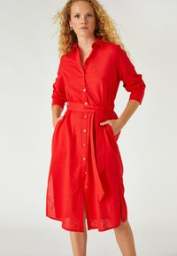 Vestido rojo de lino con botones en la parte delantera, mangas largas, un cinturón atado en la cintura y bolsillos laterales. Corte suelto con un dobladillo a la altura de la rodilla.