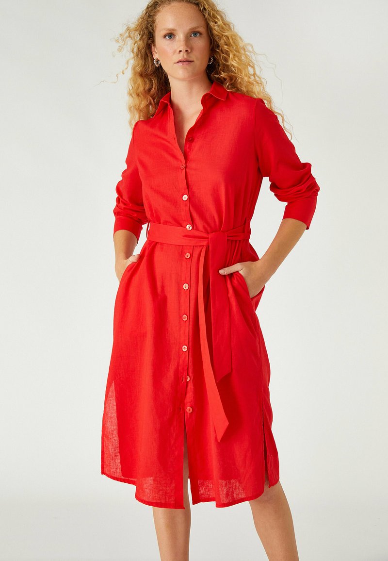 Vestido rojo de lino con botones en la parte delantera, mangas largas, un cinturón atado en la cintura y bolsillos laterales. Corte suelto con un dobladillo a la altura de la rodilla.