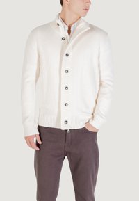 Hombre con un cárdigan blanco abotonado sobre una camisa blanca y pantalones marrón oscuro, con una mano en el bolsillo, mirando hacia un lado.