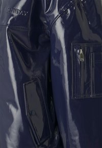 Námornícka modrá, lesklá, lakovaná látková bunda so zipsovou kapsou a embosovaným logom. Má elegantné línie a hladkú textúru.