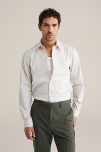 Hvid skjorte med knapper og en afslappet pasform, kombineret med skræddersyede bukser i mørkegrøn, har en glat tekstur og folder ved halsen.