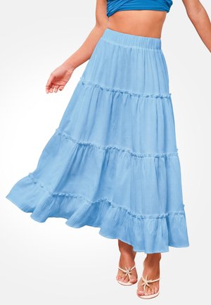 Donna che indossa una gonna lunga azzurro chiaro a balze fluente con orli arricciati e sandali bianchi decorati con stelle marine.